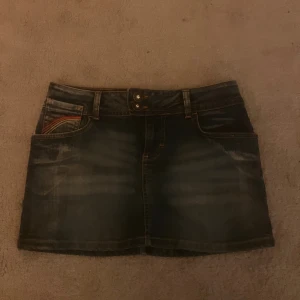 Mörk jeanskjol med regnbågsdetaljer - Snygg mörkblå jeanskjol från Love Label med regnbågsfärgade sömmar på fickorna och dubbla knappar med glittriga stenar i midjan. Klassisk femficksmodell, bälteshällor och dragkedja. Perfekt för dig som gillar denim med en twist.