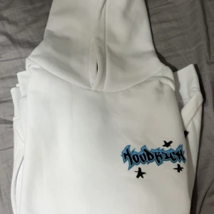 Vit hoodie från Hoodrich med tryck - Säljer en vit hoodie från Hoodrich med blå och svarta graffiti-inspirerade tryck både fram och bak. Tröjan har huva, magficka och långa ärmar. Perfekt för dig som gillar streetwear och vill sticka ut med coola detaljer.