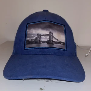 Blå keps från Gårda med Londonmotiv - Snygg blå keps från Gårda med mesh på baksidan och justerbar snapback. Framtill finns ett svartvitt motiv av Tower Bridge i London, inramat med blå kant. Perfekt för dig som gillar coola prints och streetstyle.