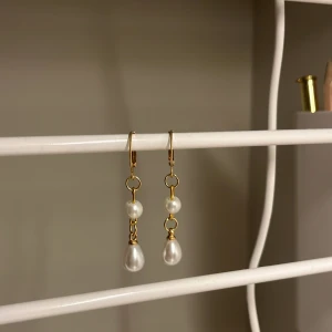 Guldörhängen med pärlor - Snygga örhängen i guldton med två vita pärlor på varje, en rund och en droppformad. Hängande design som ger en elegant vibe och passar perfekt till din stil. Perfekt accessoar för att lyfta din look. Rostfritt stål och handgjort 
