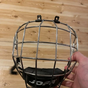 Jofa 480M hockeygaller i silver - Säljer ett Jofa 480M hockeygaller i silverfärgat metall med svart hakskydd. Gallret har robust konstruktion och fästen upptill för enkel montering på hjälm. Perfekt för dig som vill ha extra skydd på isen och gillar klassisk hockeystil.