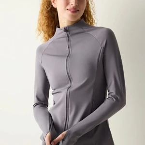 Grå vindjacka med dragkedja - Snygg grå sportjacka med hel dragkedja framtill och hög krage. Jackan har långärmade ärmar med tumhål och är tillverkad i ett mjukt, stretchigt syntetmaterial som passar perfekt till träning. Enkel och stilren design utan mönster.