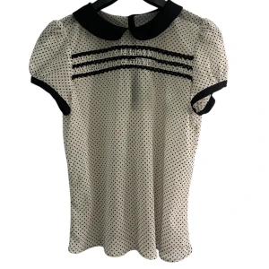 Prickig blus med svart krage - Supersöt blus i vitt med svarta prickar och kontrasterande svart krage. Puffiga korta ärmar med svarta kanter och dekorativa knappar bak. Blusen har fina spetsdetaljer framtill och är lätt och lite genomskinlig i materialet.