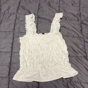 Vit rynkad linnetopp med volang - Supersöt vit linnetopp med breda axelband och rynkade detaljer över hela toppen. Volangkant nertill och små broderade hål ger en romantisk vibe. Tyvärr så är inte prislappen kvar med den är köpt på h&m och är storlek xs