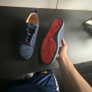 Blå sneakers från Christian Louboutin - Säljer ett par snygga blå sneakers från Christian Louboutin med röd signatursula och snörning. Skorna är tillverkade i mocka och har en rund tå samt platt sula. Insidan är beige och ger en lyxig känsla. Perfekt för dig som vill sticka ut med exklusiva detaljer.