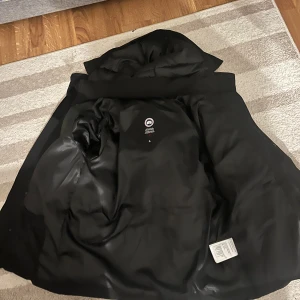 Svart dunjacka från Canada Goose - Säljer en svart dunjacka från Canada Goose med klassisk logga på ärmen. Jackan har en puffig design, huva och långa ärmar. Materialet är slitstark polyester och dunfyllning som håller dig varm under kalla dagar. Perfekt för dig som vill ha en stilren och funktionell vinterjacka.