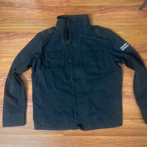 Svart parkajacka Admitted & Feeling M - Svart parkajacka från Admitted & Feeling med hög krage, flera fickor med lock och knappstängning, samt broderad logga på ärmen. Jackan har både dragkedja och knappar framtill och är tillverkad i bomull. Snygga axeldetaljer med knappar ger en cool look.