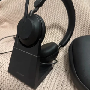 Jabra Evolve2 65 med laddningsställ - Jabra Evolve2 65 trådlöst headset med laddningsställ och USB-A, optimerat för Microsoft Teams. Svart färg, med tillhörande hårt fodral. Headsetet ser ut att vara i mycket gott skick utan synligt slitage eller defekter.