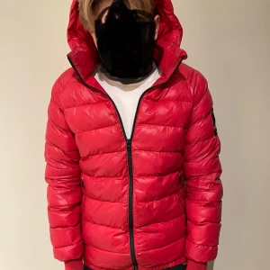 Röd Peak Performance dunjacka 170 - Snygg röd dunjacka från Peak Performance med huva och svart logotyp på ärmen. Jackan har quiltad design, dragkedja framtill och två sidofickor med dragkedja. Perfekt för kalla dagar och har en modern, sportig look som passar dig som gillar streetstyle. Den är lite sönder i vänstra fickan.