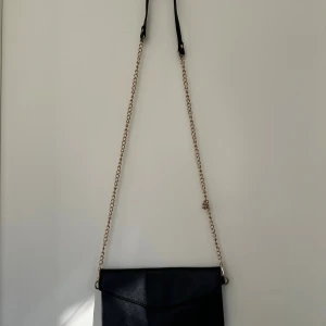 Svart kuvertväska med guldkedja - Stilren svart kuvertväska med guldfärgad kedja som axelrem. Väskan har ett magnetlås och insidan är fodrad med mönstrat tyg och har en innerficka med dragkedja. Perfekt för att bära det viktigaste och ge en snygg touch till din outfit.