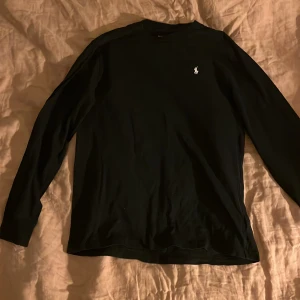 Svart långärmad tröja Polo Ralph Lauren - Svart långärmad tröja från Polo Ralph Lauren med klassisk vit logobrodyr på bröstet. Tröjan har rund hals och är gjord i mjuk bomull, perfekt för chill dagar. Enkel och stilren design som funkar till allt.