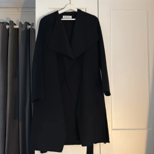 Svart kappa från Wera med bälte - Svart kappa från Wera i stilren design bälte i midjan. Snygg omlott-look framtill. Perfekt för dig som gillar minimalistisk och klassisk stil. Materialet är mjukt och känns lyxigt. Inga anmärkningar på kappan och endast använd ett fåtal gånger. Som ny