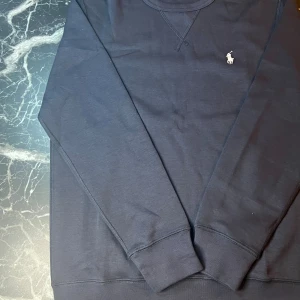 Mörkblå tröja från Polo Ralph Lauren - Snygg mörkblå långärmad tröja från Polo Ralph Lauren med klassisk rund halsringning och broderad vit logga på bröstet. Tillverkad i mjuk bomull som känns skön mot huden. Perfekt för dig som gillar stilren och enkel design.