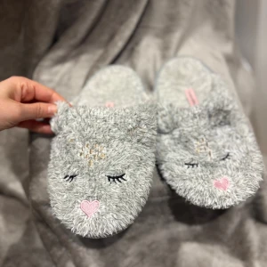 Grå fluffiga tofflor från Hunkemöller - Supergulliga grå tofflor från Hunkemöller med broderat djuransikte, stängda ögon och rosa hjärta på tån. De är gjorda i mjukt, fluffigt syntetmaterial och har små öron upptill. Perfekta för att mysa runt hemma under kalla dagar.