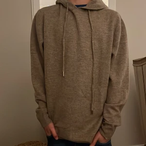 Beige hoodie i merinoull - Säljer en beige hoodie med huva och långa ärmar. Tröjan har snörning vid halsen och är tillverkad i ett mjukt material som känns skönt mot huden. Perfekt för chill dagar och enkel att matcha med jeans eller joggers. I merinoull och 20% cashmere