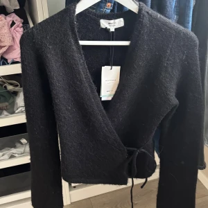 Svart omlottkofta från Gina Tricot - Svart omlottkofta i mjuk ullblandning från Gina Tricot. Koftan har långa ärmar, knytband i midjan och en snygg v-ringad design. Snöret är i den tunnare varianten, helt ny aldrig använd🥰skriv privat vid frågor 