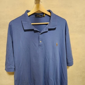 Blå pikétröja Polo Ralph Lauren - Snygg blå pikétröja från Polo Ralph Lauren i storlek XLT. Klassisk krage, korta ärmar och två knappar framtill. Tröjan har det ikoniska broderade polospelaren på bröstet. Tillverkad i mjuk bomull som känns skön mot huden.
