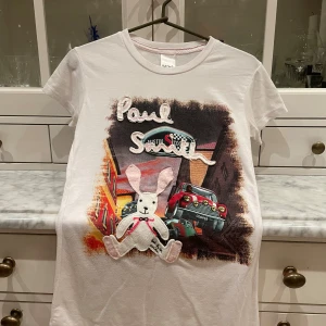 Vit Paul Smith t-shirt med kanin och bil - Vit t-shirt från Paul Smith med coolt tryck av bilar och stadsmiljö på framsidan. Broderad logga och en söt 3D-kanin i tyg framtill. Klassisk passform med korta ärmar och rund hals. Perfekt för dig som gillar unika detaljer och streetstyle.    Observera att det är sytt på ett ”slarvigt” sett med lösa trådar etc. Storlek visar L men är som small, fungerar både som lösare modell el tight 
