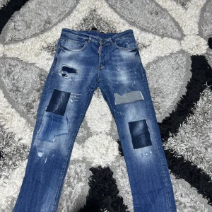 Patchade blå jeans från Dsquared2 - Patchade blå jeans från Dsquared2 i storlek 48. Snyggt slitna med flera olika tygpatchar och rivna detaljer framtill. Klassisk fyrficksmodell med knappgylf och cool tvättad look. Perfekt för dig som gillar streetstyle och unika plagg.
