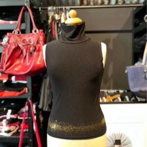 Svart polotopp med guldglitter från Koola Anna - Svart ärmlös polotopp från Koola Anna i stretchigt polyester-material. Toppen har en hög krage och är dekorerad med guldglittrande detaljer längst ner. Snygg och stilren modell som ger en cool vibe med sin glittriga finish.