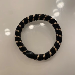 Svart och guldigt armband från KENKKI - Snyggt armband från KENKKI i svart och guld. Armbandet är flätat med ett grovt band och har inslag av glittrig guldtråd som ger en cool kontrast. Det har en rund form och en svart plastdetalj med varumärket ingraverat.