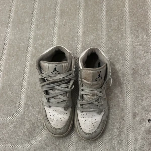 Nike Air Jordan 1 vita sneakers - Nike Air Jordan 1 sneakers i vitt med grå detaljer och klassisk Jordan-logga på plösen. Skorna har snörning, perforerad tå och platt sula. Tillverkade i skinn med en rund tå och högt skaft. Perfekt för dig som gillar streetstyle och ikoniska sneakers.