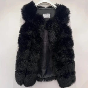 Svart fluffig fuskpäls väst från Lovely - Svart väst från Lovely med superfluffig och mjuk känsla. Västen har en rak passform och är fodrad på insidan för extra komfort. Perfekt att slänga över en hoodie eller långärmad tröja för en cool look. Snygg och stilren design utan krusiduller.