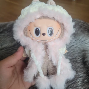 Fluffig vit och rosa kappa till Labubu  - Supergullig kappa i fluffigt vitt garn med ljusrosa fuskpäls runt kanten. Huvan har snören med fluffiga bollar i ändarna och är stickad för extra mysig känsla. 