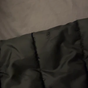 Svart pufferjacka från 2117 - Svart pufferjacka från 2117 med huva och dragkedja framtill. Jackan har quiltad design och två sidofickor med dragkedja. Materialet är syntet och jackan har en stilren look med vitt tryck på bröstet. Perfekt för kyliga dagar. Har dock ett jätte litet hål på ärmen