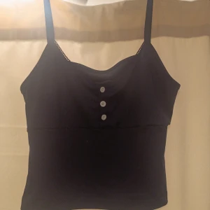 Marinblå linnetopp med knappar - Marinblå linnetopp med smala axelband och tre vita knappar framtill. Toppen har en enkel och stilren design med lätt rundad halsringning. Materialet känns mjukt och bekvämt, perfekt för varma dagar. Snygg detalj med knapparna som ger lite extra karaktär!❤️