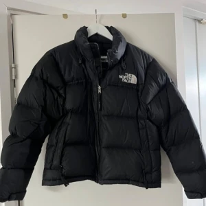 Svart pufferjacka från The North Face - Säljer en svart pufferjacka från The North Face med hög krage och dragkedja framtill. Jackan har en klassisk boxig passform och är perfekt för kalla dagar. Ikonisk logga på bröstet och vadderad design för extra värme.