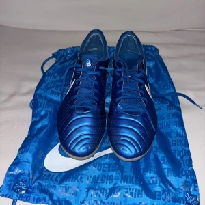 Blå Nike Tiempo fotbollsskor - Säljer ett par Nike Tiempo fotbollsskor i metallicblå med svarta och vita detaljer. Skorna har snörning, rund tå och platt sula med dobbar för gräsplan. Ovandelen är tillverkad i syntet och skinn för bästa känsla och kontroll. Snygg design med stora vita swoosh-loggor på sidorna.