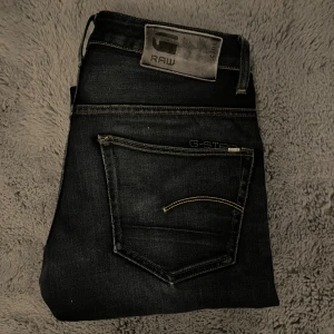 G-Star RAW 3301 - Mörkblåa G-Star RAW 3301 jeans med straight fit och slitna detaljer på knä och lår. Klassisk femficksmodell med kontrastsömmar och RAW-logga på bakfickan. Snyggt tvättad denim som ger en cool look. Storlek 30/32