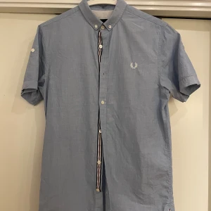 Blå kortärmad skjorta Fred Perry - Snygg ljusblå kortärmad skjorta från Fred Perry i slim fit-modell. Skjortan har klassisk button down-krage, vita knappar och broderad logga på bröstet med unik design. Tillverkad i bomull och perfekt för en clean och stilren look.
