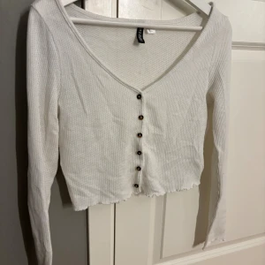 Vit croppad topp med knappar H&M - Säljer en vit croppad topp från H&M Divided i storlek M. Toppen har lång ärm, v-ringning och snygga bruna knappar framtill. Materialet är ribbad bomull som ger en skön och avslappnad känsla. Perfekt för dig som gillar enkel och stilren look.