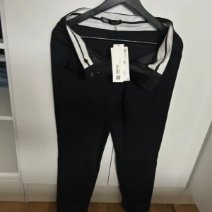 Svarta kostymbyxor från Zara - Snygga svarta kostymbyxor från Zara med klassisk passform och diskret knappstängning. Byxorna har en stilren look och är perfekta för dig som gillar en clean och dressad stil. Materialet känns mjukt och följsamt.