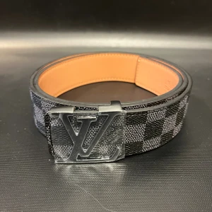 Svart rutig skinnbälte med silver logga - Säljer ett svart bälte med rutigt mönster i svart och grått, bred design med en silver metall logga på spännet. Insidan är i ljusbrunt skinn med präglad märkning. Perfekt för dig som vill ha en lyxig och stilren accessoar med statement-känsla.