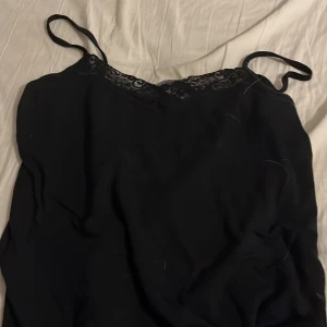Svart linnetopp med spets från 157 - Svart linnetopp från 157 med tunna axelband och snygg spetsdetalj längs urringningen. Enkel och stilren modell som är perfekt att matcha med jeans eller kjol. Toppens material är jättemjukt och de är storlek S