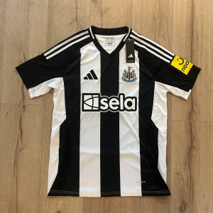 Newcastle United Isak 14 Adidas tröja - Säljer en officiell Newcastle United matchtröja från Adidas med Isak 14 på ryggen. Tröjan är svart och vit med breda ränder, korta ärmar och V-ringad krage. Materialet är lätt polyester med Aeroready-teknologi. Klubbmärket och sponsorlogga på bröstet.