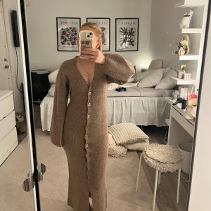 Adoore från Ginatricot - Mysig beige stickad långklänning med v-ringning och stora knappar hela vägen framtill. Klänningen har långa ärmar och en figurnära passform som ändå känns avslappnad. Perfekt för dig som gillar en enkel men trendig look. Helt slutsåld på nätet hos gina