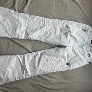 Vita täckbyxor från AST XS - Säljer ett par vita täckbyxor från AST i storlek XS. Byxorna har justerbara hängslen, dragkedja och knapp framtill samt flera fickor. Materialet är vattentätt och andas, perfekt för kalla dagar. Snygg och clean look med detaljer i svart.