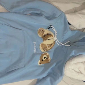 Blå Palm Angels hoodie med nalle - Säljer en ljusblå hoodie från Palm Angels med broderad nallebjörn på bröstet. Tröjan har huva med snörning och en stor magficka. Ikonisk Palm Angels-logga i vitt under motivet. Perfekt för dig som gillar streetwear och unika detaljer.