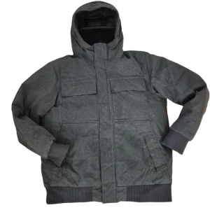 Grå dunjacka från The North Face - Säljer en grå dunjacka från The North Face med huva, ribbade muddar och flera praktiska fickor. Jackan har broderad logga på bröstet och stängs med dragkedja och knappar. Perfekt för kalla dagar när du vill hålla värmen och ändå se stilren ut.