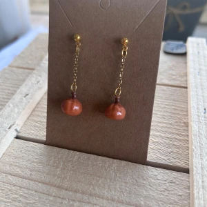 Guldiga örhängen med orange pumpa hängen - Unika örhängen med tunn guldfärgad kedja och små, runda orange hängen som påminner om minipumpor. Fästs med guldfärgade pluppar och har en lekfull, handgjord känsla. Perfekta för dig som vill sticka ut med färg och form.