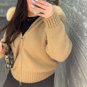 Beige stickad jacka med pälshuva alessa ÄKTA PÄLS - Säljer en beige stickad jacka med fluffig pälskant på huvan från Alessa. Jackan har dragkedja framtill, ribbade muddar och en loose passform. Perfekt för dig som vill ha en mysig och snygg look med extra fluff på huvan. Den är använd 2 gånger och som ny. 