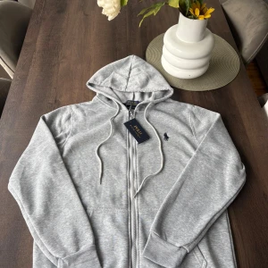 Grå hoodie från Polo Ralph Lauren - Snygg ljusgrå hoodie från Polo Ralph Lauren med dragkedja och broderad marinblå logga på bröstet. Slim fit-modell med huva och snörning, tillverkad i mjuk bomullsblandning. Perfekt för en chill och stilren look.