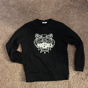 Svart Kenzo sweatshirt med tigertryck - Svart sweatshirt från Kenzo med ikoniskt tigertryck och logga framtill. Tröjan har rund halsringning, långa ärmar och ribbade muddar. Perfekt för dig som vill sticka ut med en klassisk streetstyle-look.