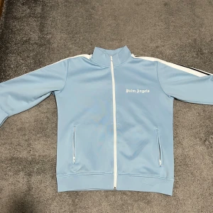 Ljusblå Palm Angels - Säljer en ljusblå  Palm Angels med vit dragkedja och vita ränder längs axlarna. Den har ståkrage, två fickor med dragkedja och logotyp på bröstet. Materialet är polyester och passformen är normal. Jag har använt den några gånger och har inte haft intresse för den. Priset kan diskuteras.
