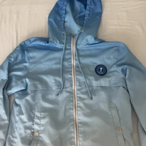 Blå vindjacka från Trapstar med huva - Snygg ljusblå vindjacka från Trapstar med broderad logga på bröstet och stor Trapstar-text på ryggen. Jackan har huva med dragsko, hel dragkedja framtill och två fickor med knappar. Perfekt för dig som vill sticka ut med streetwear-vibe.