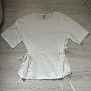 Vit blus med knyt i sidan - Säljer en vit blus med rund hals och korta ärmar. Toppen har snygga knytband i båda sidorna som ger en cool detalj och lite form till midjan. Materialet är mjukt och känns som bomull, perfekt för vår och sommar. Enkel och stilren design som funkar till många outfits. Aldrig använd 